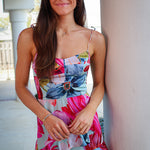 Woman wearing a colorful floral dress and denim shorts standing against a light-colored wall.
