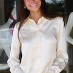 A smiling woman with long dark hair wearing a cream-colored satin button-down shirt and blue jeans, standing in front of a glass storefront.