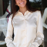 A smiling woman with long dark hair wearing a cream-colored satin button-down shirt and blue jeans, standing in front of a glass storefront.