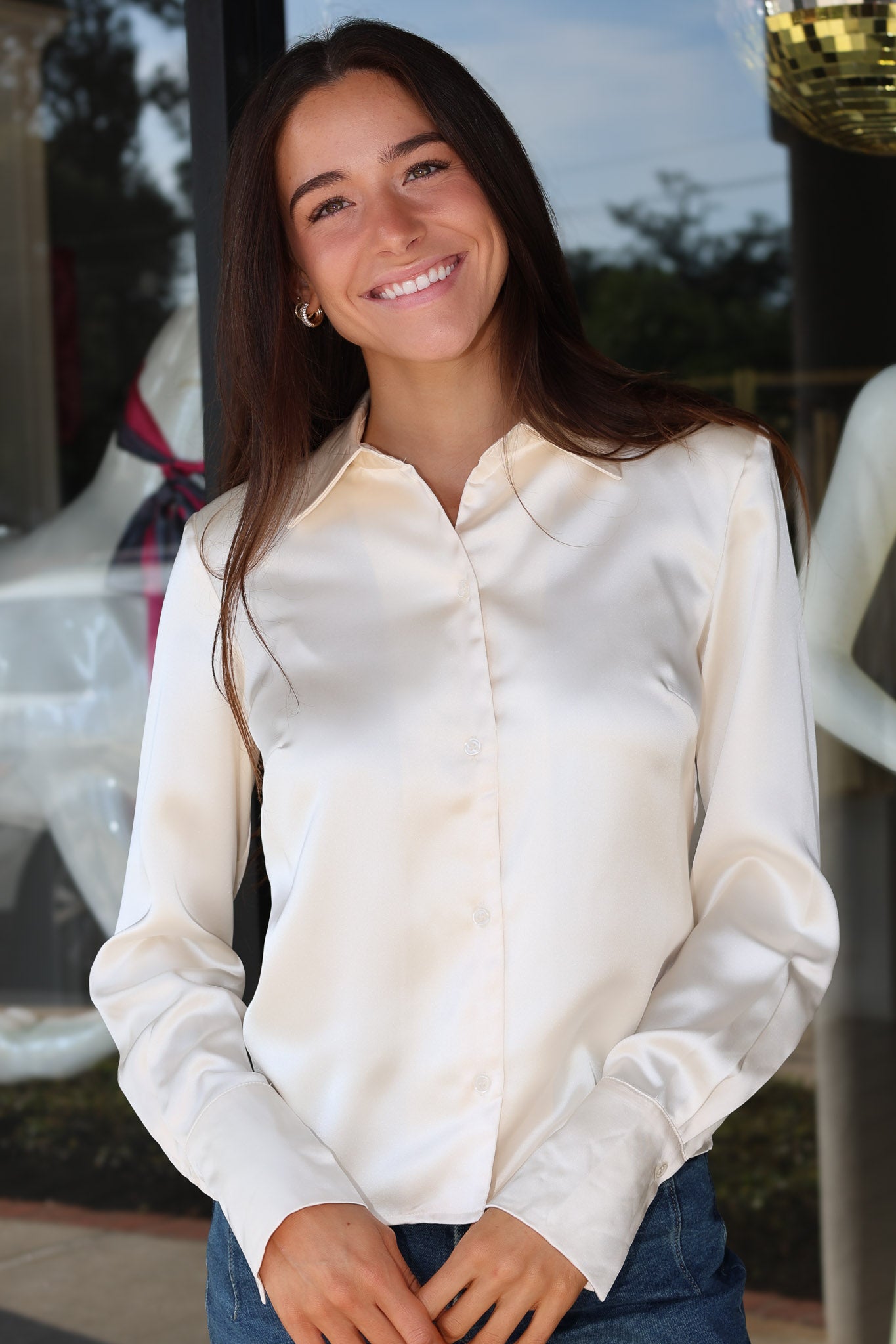 A smiling woman with long dark hair wearing a cream-colored satin button-down shirt and blue jeans, standing in front of a glass storefront.