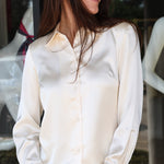 A woman wearing a long-sleeve cream satin button-up shirt and blue jeans, standing in front of a storefront window.