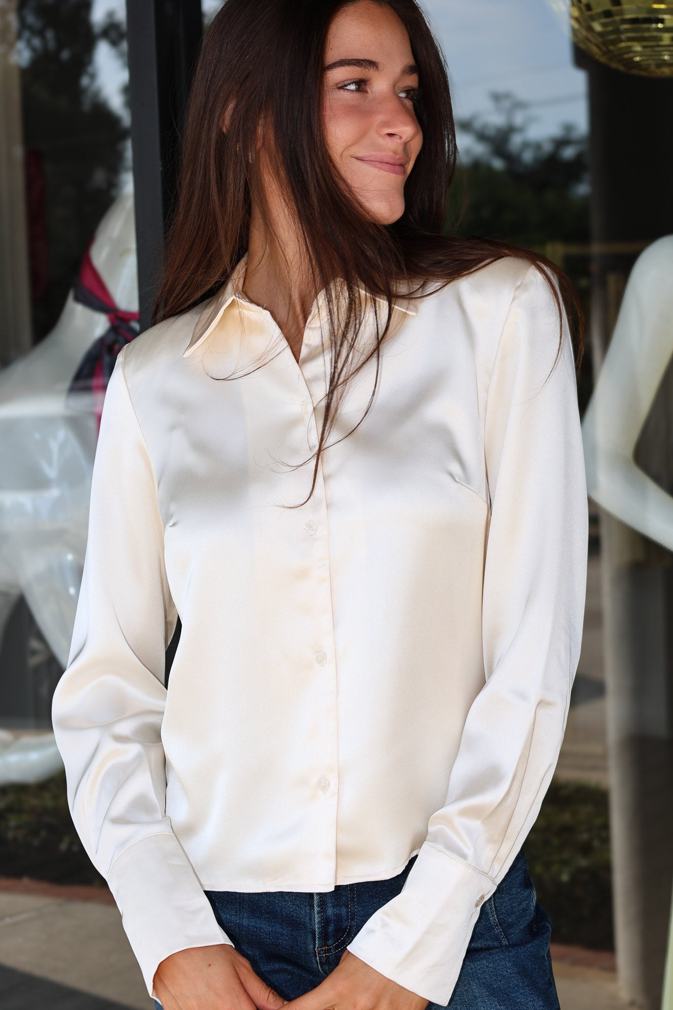 A woman wearing a long-sleeve cream satin button-up shirt and blue jeans, standing in front of a storefront window.