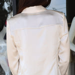 A woman viewed from behind wearing a light cream satin button down and blue jeans, standing in front of a glass storefront.