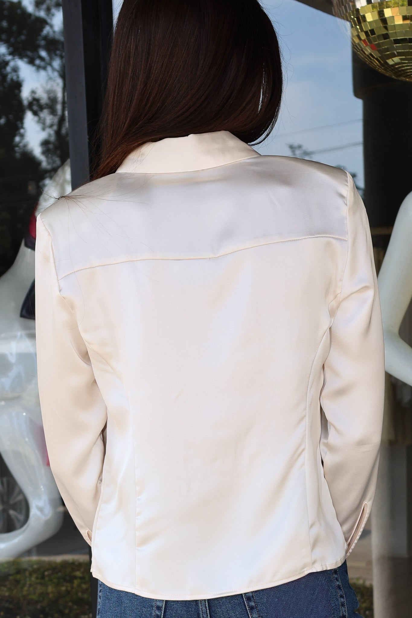 A woman viewed from behind wearing a light cream satin button down and blue jeans, standing in front of a glass storefront.