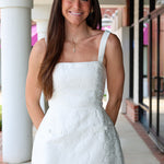 Woman in a white jaquard floral dress standing outdoors near a building