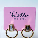 A pair of gold-tone ring earrings with rectangular clear gemstones, displayed on a pink card labeled 'Rodéo Boutique' against a light blue-to-white gradient background.