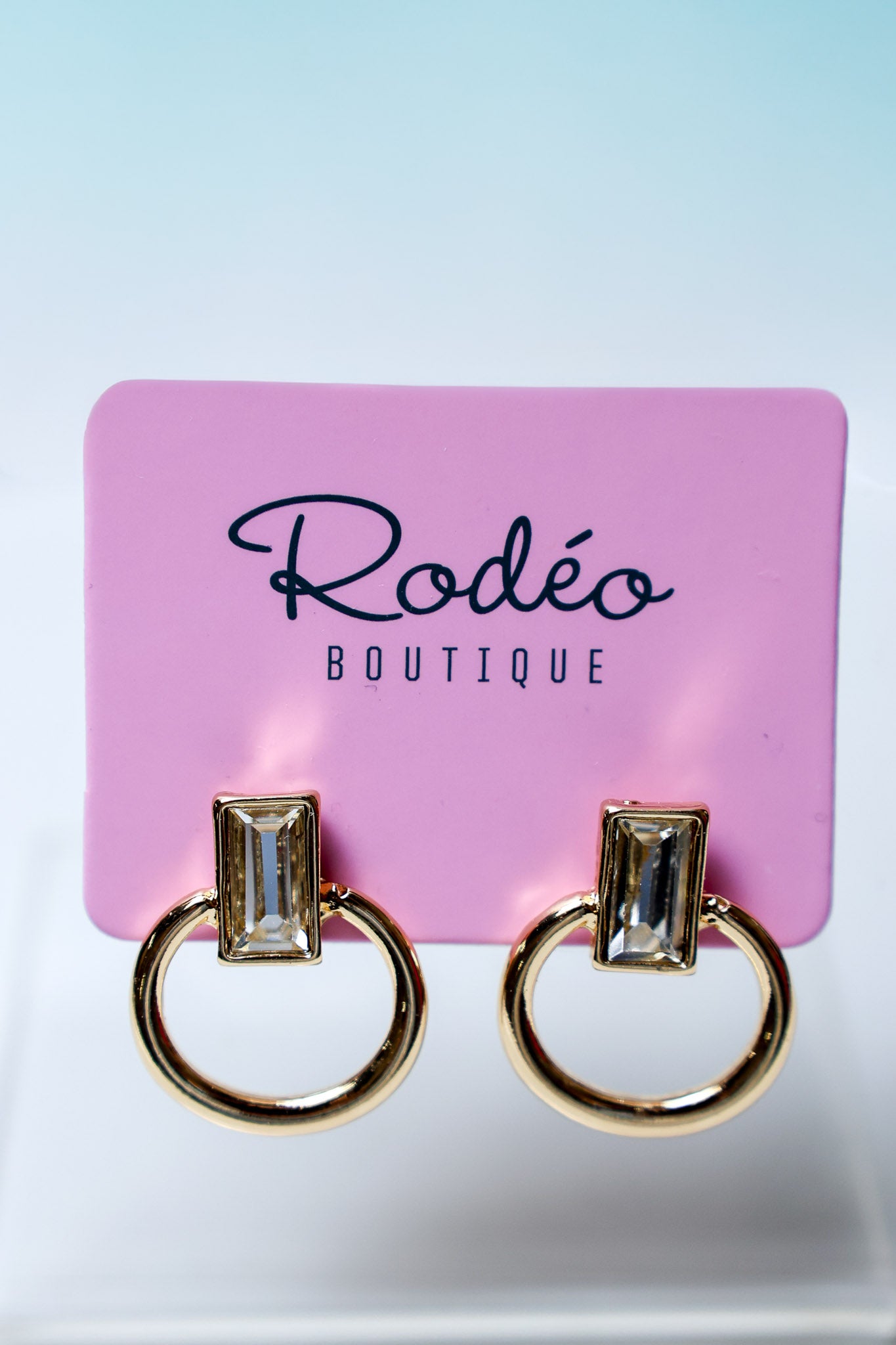 A pair of gold-tone ring earrings with rectangular clear gemstones, displayed on a pink card labeled 'Rodéo Boutique' against a light blue-to-white gradient background.