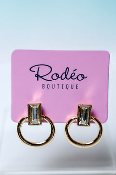 A pair of gold-tone ring earrings with rectangular clear gemstones, displayed on a pink card labeled 'Rodéo Boutique' against a light blue-to-white gradient background.