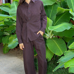 A woman wearing a matching dark brown and black checkered long-sleeve button-up shirt and wide-leg pants set, standing outdoors in front of large green tropical leaves.