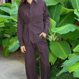 A woman wearing a matching dark brown and black checkered long-sleeve button-up shirt and wide-leg pants set, standing outdoors in front of large green tropical leaves.