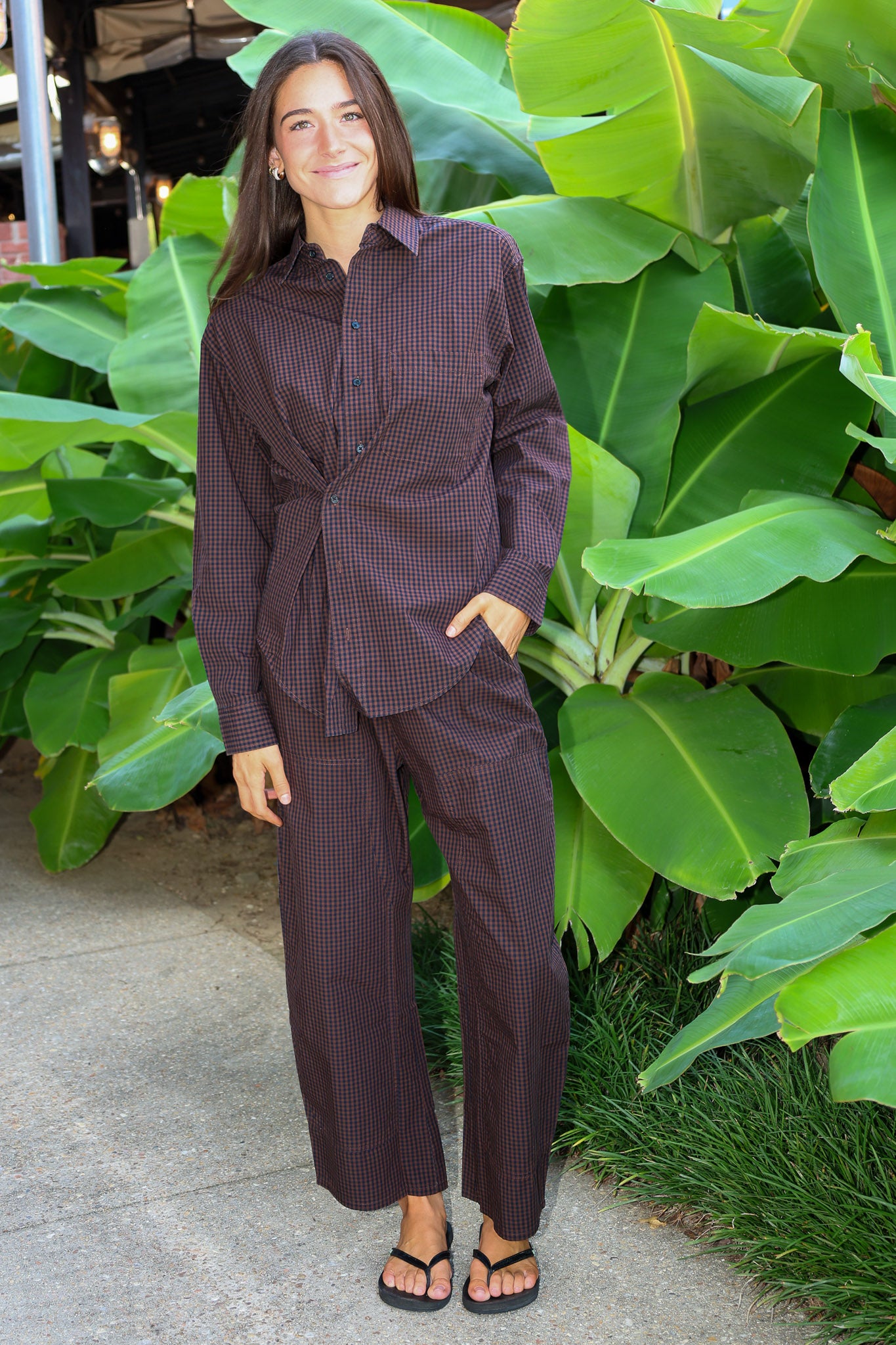 A woman wearing a matching dark brown and black checkered long-sleeve button-up shirt and wide-leg pants set, standing outdoors in front of large green tropical leaves.