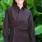 A woman smiling in front of large green tropical leaves, wearing a dark brown and black checkered button-down shirt with a matching dark skirt or pants.