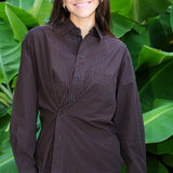 A woman smiling in front of large green tropical leaves, wearing a dark brown and black checkered button-down shirt with a matching dark skirt or pants.