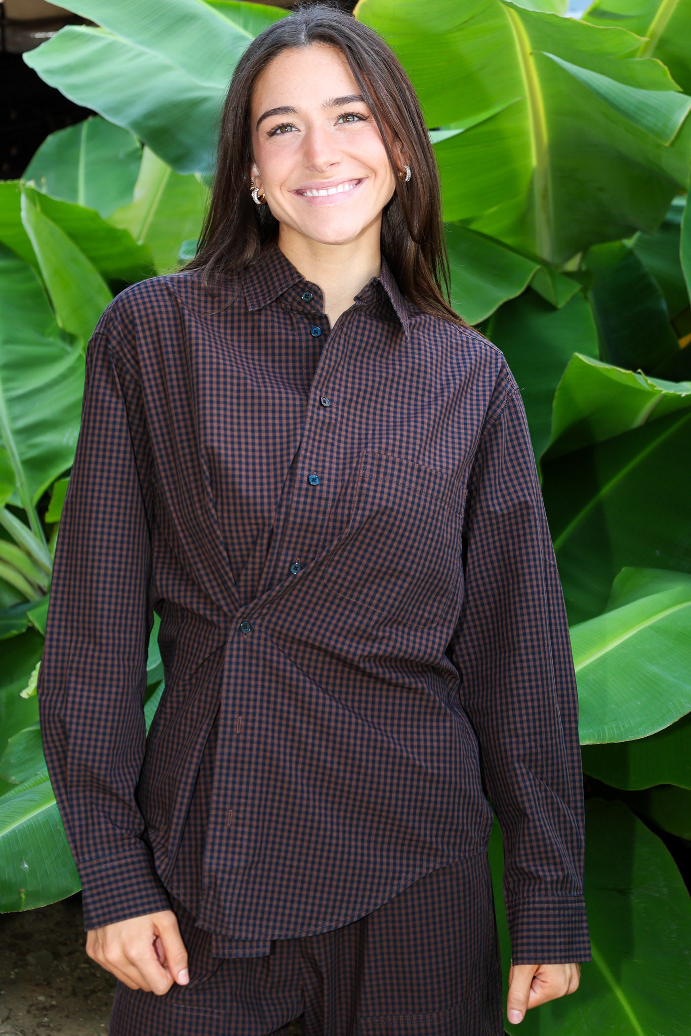 A woman smiling in front of large green tropical leaves, wearing a dark brown and black checkered button-down shirt with a matching dark skirt or pants.