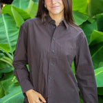 A woman wearing a dark brown and black checkered button-down shirt and matching pants, standing in front of a background of large green tropical leaves.