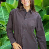 A woman wearing a dark brown and black checkered button-down shirt and matching pants, standing in front of a background of large green tropical leaves.