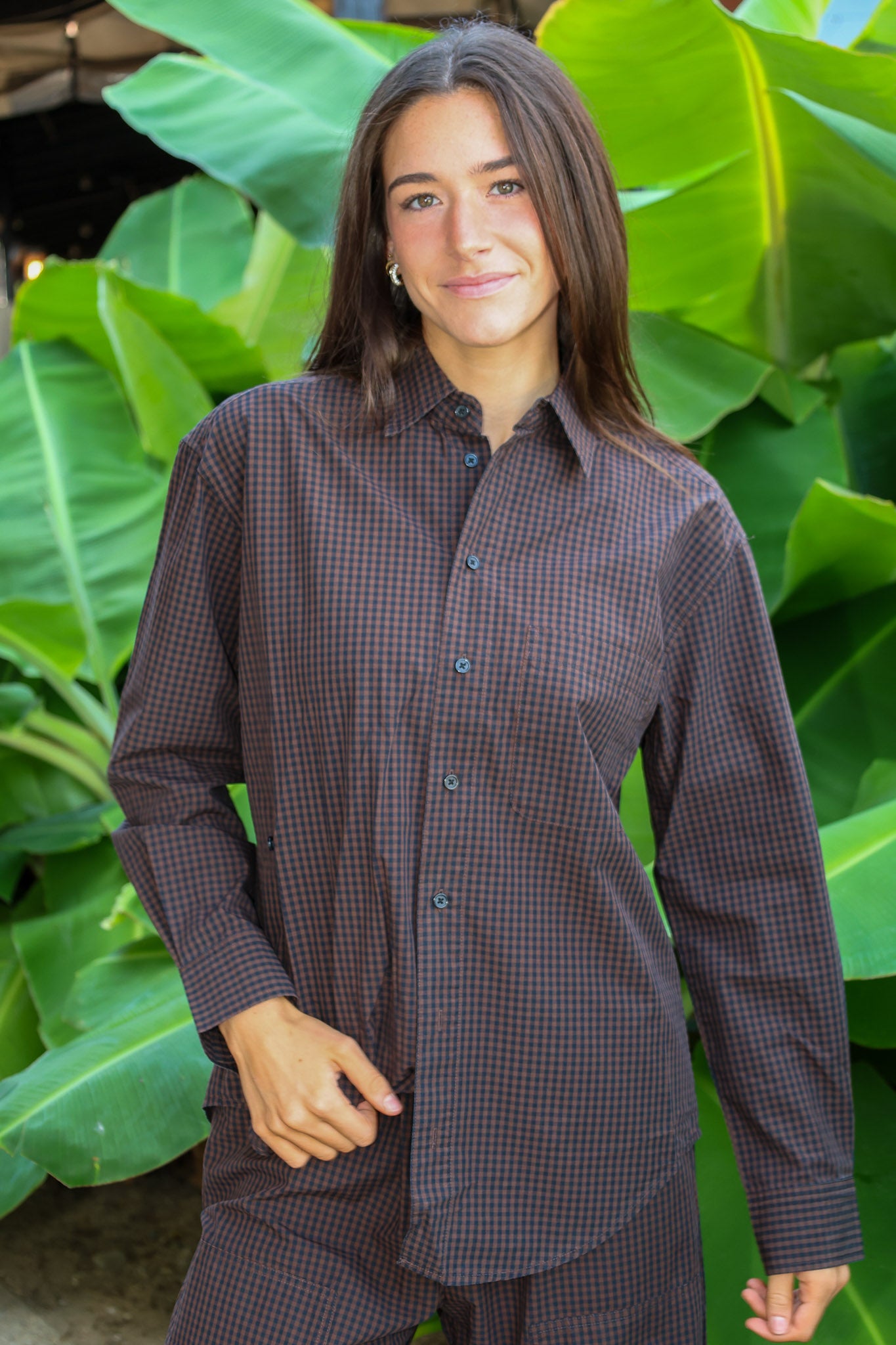 A woman wearing a dark brown and black checkered button-down shirt and matching pants, standing in front of a background of large green tropical leaves.