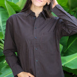 A woman wearing a dark brown and black checkered button-down shirt, posing in front of a background of large green tropical leaves.