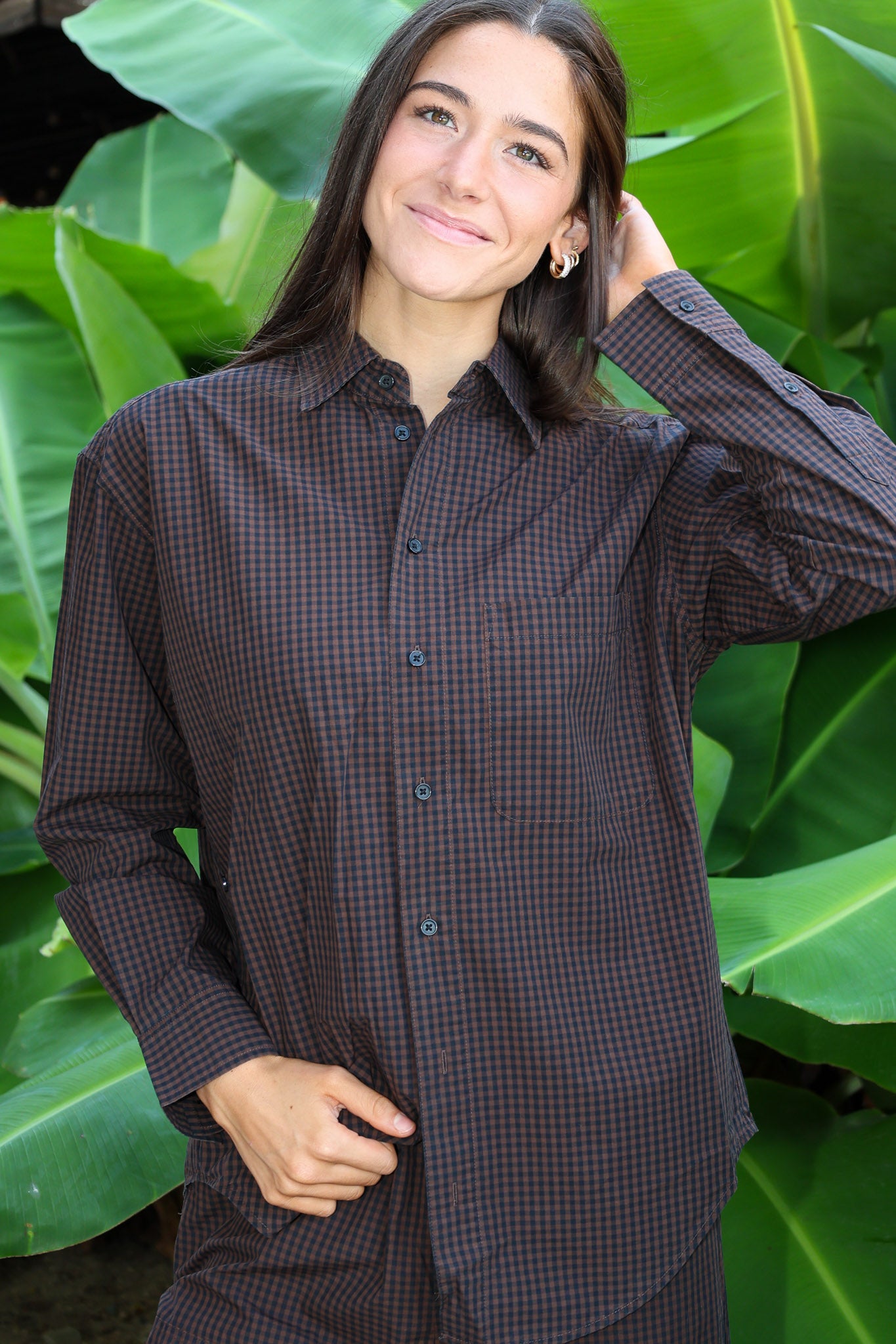 A woman wearing a dark brown and black checkered button-down shirt, posing in front of a background of large green tropical leaves.
