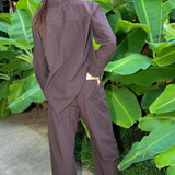 A woman wearing a brown and black micro-checkered long-sleeve shirt and matching wide-leg pants, shown from behind in an outdoor setting with large green tropical leaves.