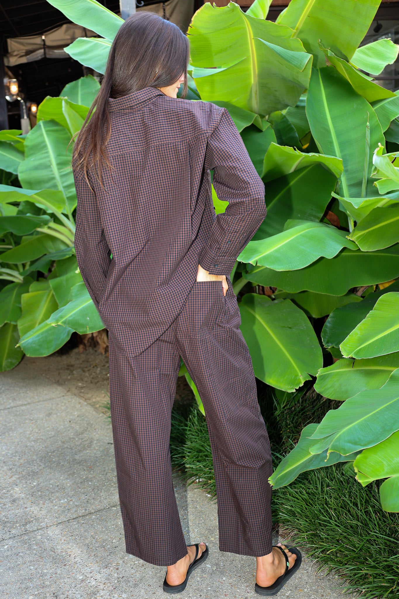A woman wearing a brown and black micro-checkered long-sleeve shirt and matching wide-leg pants, shown from behind in an outdoor setting with large green tropical leaves.