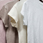 Four t-shirts in pink, gray, beige, and white hanging on a rack.