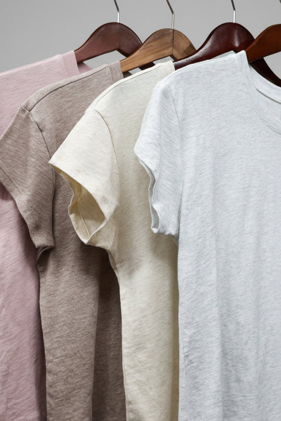 Four t-shirts in pink, gray, beige, and white hanging on a rack.