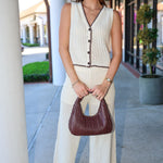 Woman in a white sleeveless crochet top and matching pants holding a brown handbag on a sidewalk.