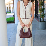Woman in a white sleeveless crochet top and matching pants holding a brown handbag on a sidewalk.