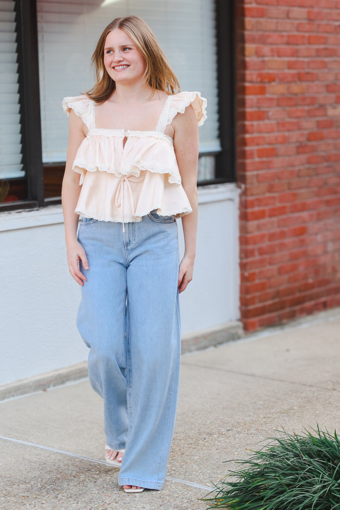 Woman wearing a ruffled top and light blue jeans standing in front of a brick building.