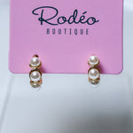 A pair of gold hoop huggie earrings with 5 stacked white pearls, displayed on a pink card labeled 'Rodéo BOUTIQUE' against a light gray background.