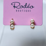 A pair of gold hoop huggie earrings with 5 stacked white pearls, displayed on a pink card labeled 'Rodéo BOUTIQUE' against a light gray background.