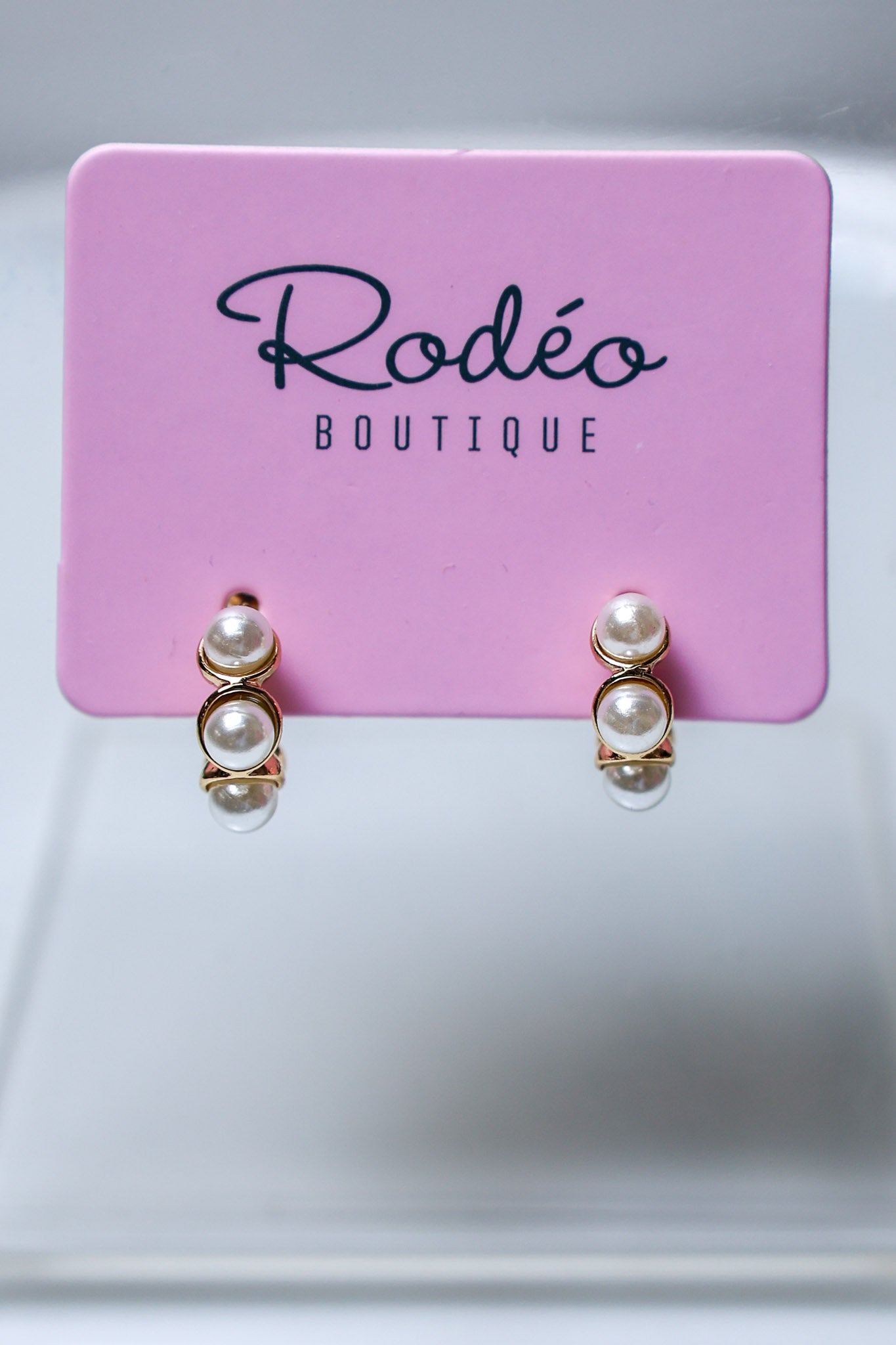 A pair of gold hoop huggie earrings with 5 stacked white pearls, displayed on a pink card labeled 'Rodéo BOUTIQUE' against a light gray background.