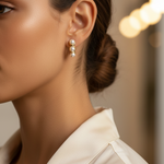 A close-up side profile of a woman wearing a delicate gold and pearl drop huggie earring,