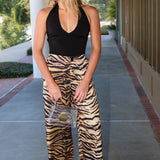 A woman stands on a covered walkway wearing a black halter top and high-waisted, wide-leg tiger-print pants in beige, brown, and black. She is holding a clear clutch bag.
