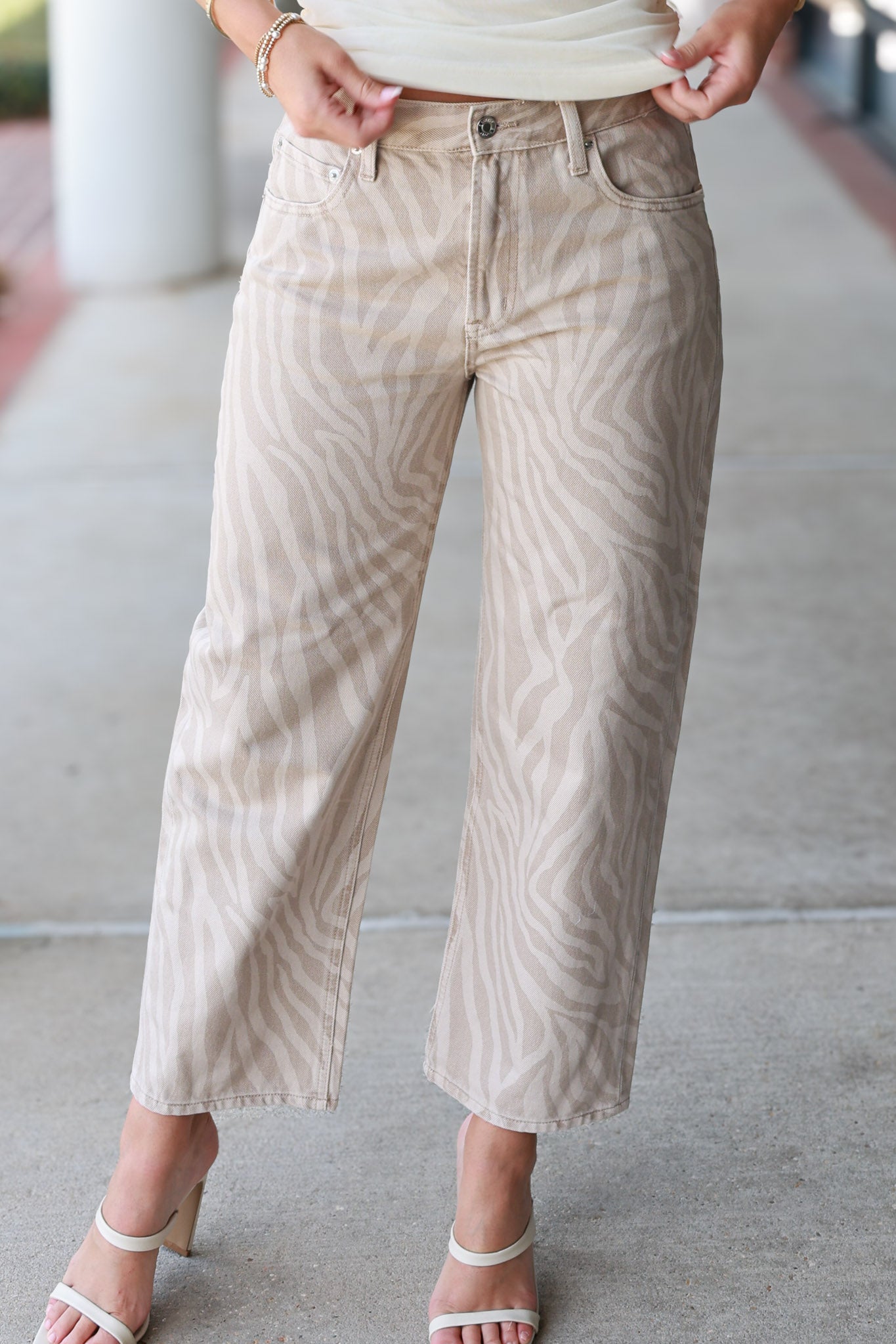 A person wearing cropped wide-leg jeans with a beige and off-white zebra-like stripe pattern, paired with white heeled sandals, standing on a concrete surface.