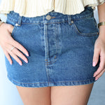 Person wearing a blue denim skirt with a white top against a light background
