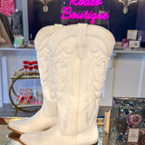A pair of off-white western cowboy boots with intricate floral embroidery on the shafts, displayed on a reflective metallic surface inside a boutique. A pink neon sign reading 'Rodeo Boutique' is visible in the background.