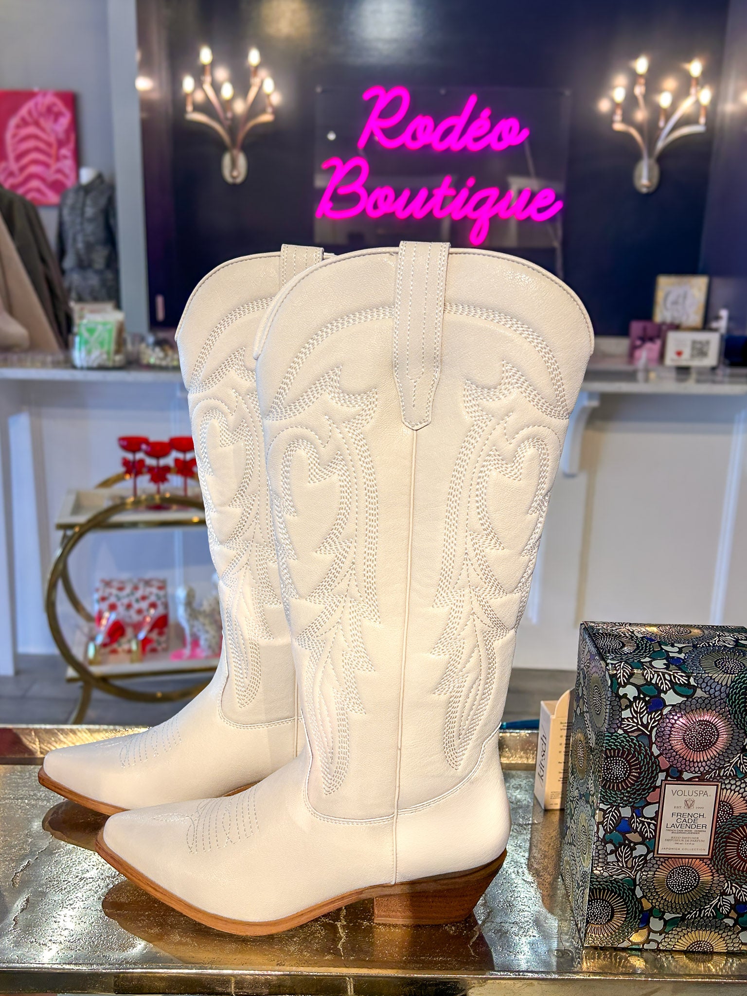 A pair of off-white western cowboy boots with intricate floral embroidery on the shafts, displayed on a reflective metallic surface inside a boutique. A pink neon sign reading 'Rodeo Boutique' is visible in the background.