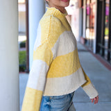 Woman wearing a yellow and white striped sweater and denim shorts standing on a sidewalk.