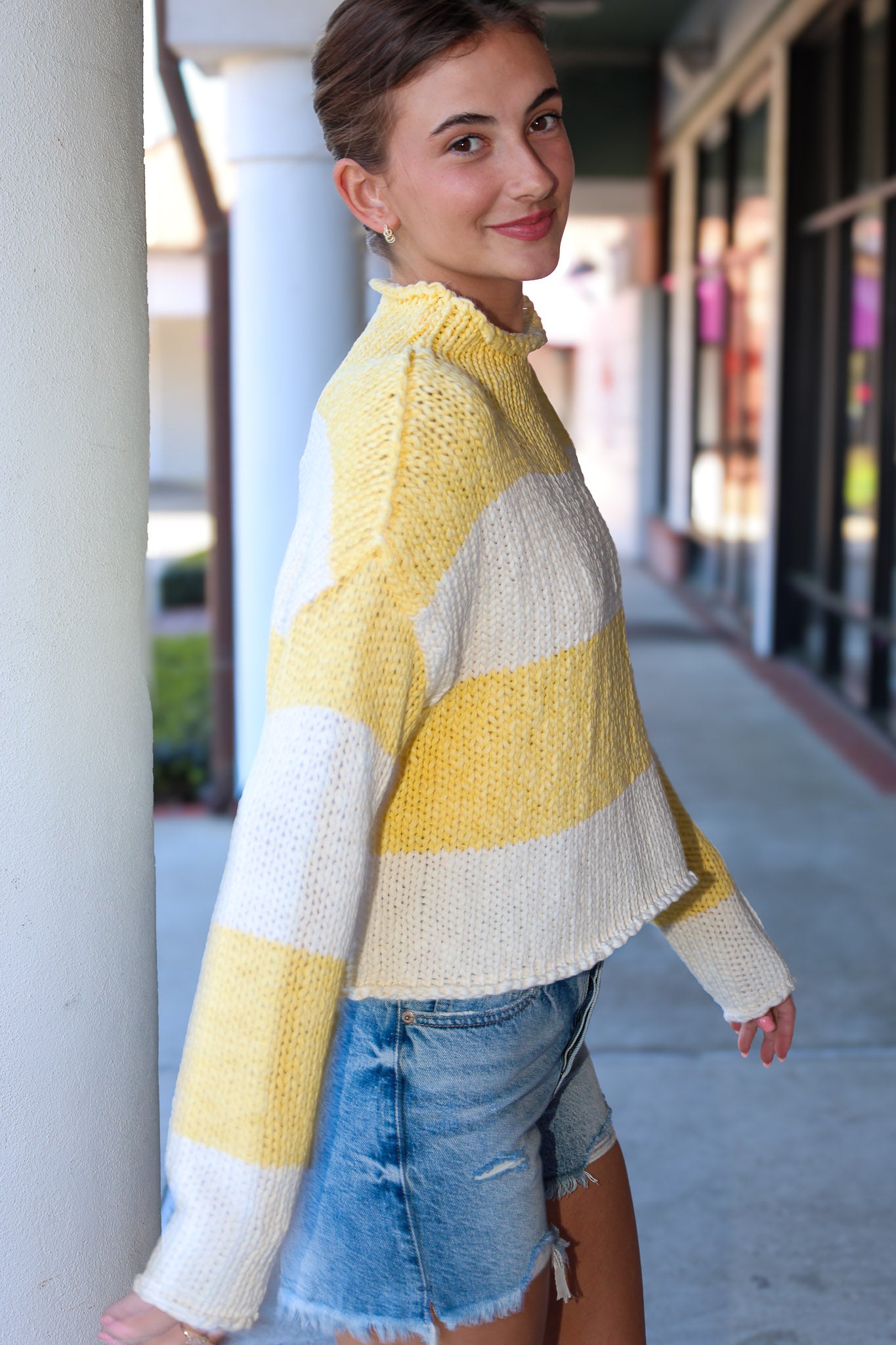 Woman wearing a yellow and white striped sweater and denim shorts standing on a sidewalk.