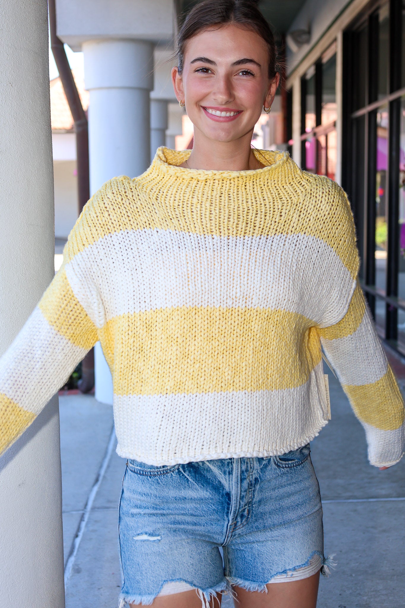 Person wearing a yellow and white striped sweater with denim shorts, standing outdoors.