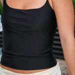 A close-up of a person wearing a black spaghetti-strap camisole top with a scoop neckline, paired with an off-white skirt. The photo is cropped from the chest down, showing the fit of the top against the model's body.