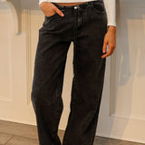 A woman wearing a white off-the-shoulder long-sleeve top and black wide-leg jeans, standing in a boutique setting with a pink neon sign in the background.