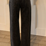 Black wide-leg denim jeans shown from the back, worn by a model standing on a wood floor in front of a white paneled wall.