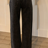 Black wide-leg denim jeans shown from the back, worn by a model standing on a wood floor in front of a white paneled wall.