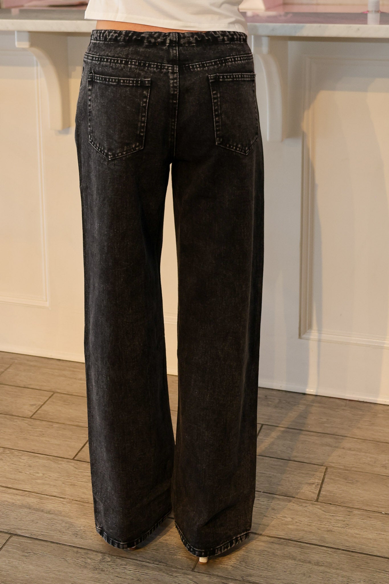 Black wide-leg denim jeans shown from the back, worn by a model standing on a wood floor in front of a white paneled wall.