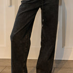 A person wearing wide-leg black denim jeans with a faded wash, paired with white strappy sandals, shown from the waist down against a white paneled wall and wood floor.