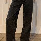 A person wearing wide-leg black denim jeans with a faded wash, paired with white strappy sandals, shown from the waist down against a white paneled wall and wood floor.