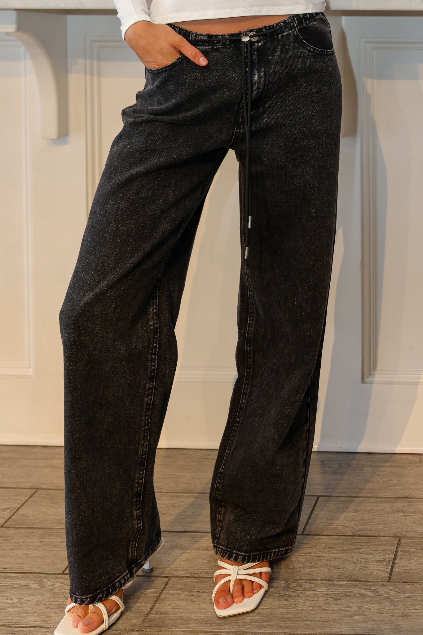 A person wearing wide-leg black denim jeans with a faded wash, paired with white strappy sandals, shown from the waist down against a white paneled wall and wood floor.